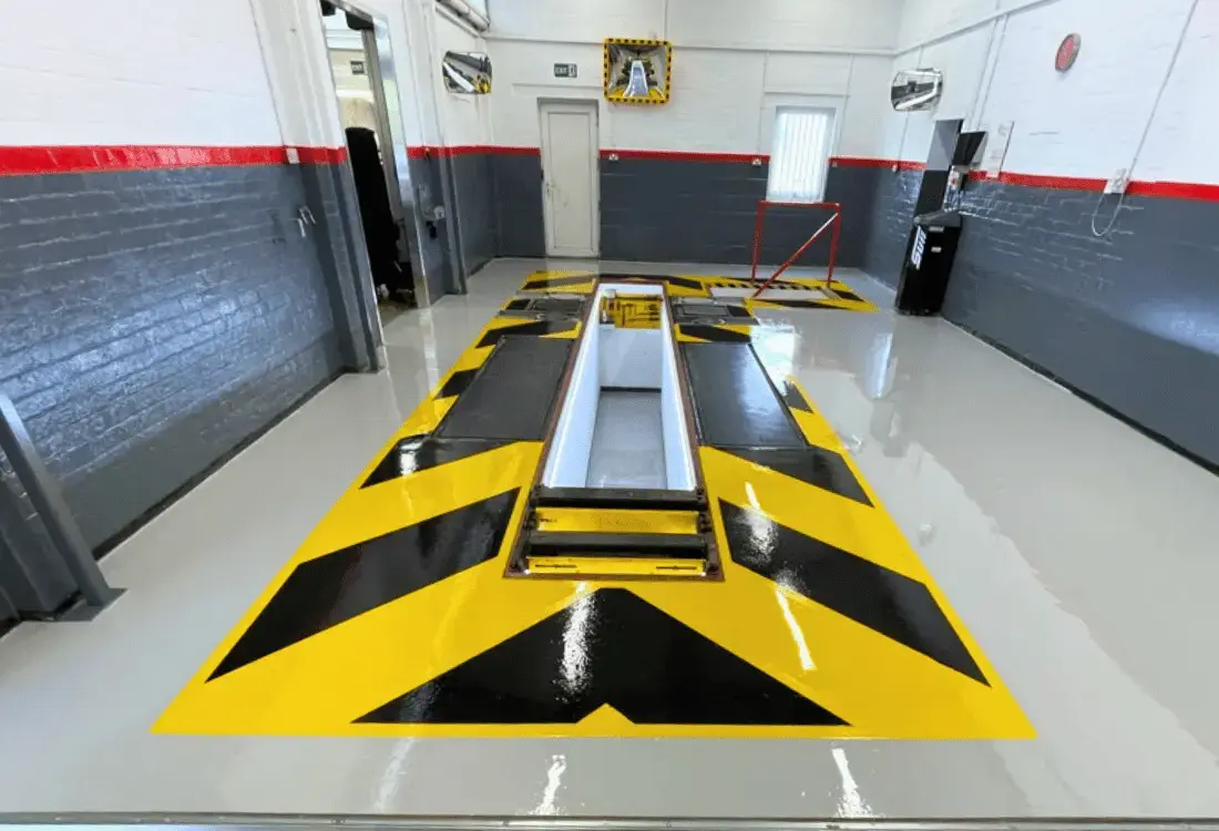 Glossy industrial resin flooring with black and yellow safety markings surrounding a vehicle inspection pit in a workshop.